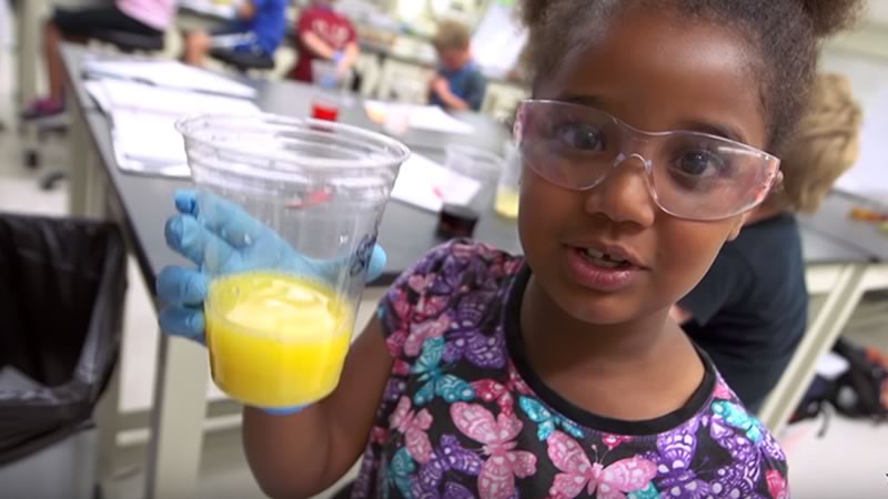 Girl wearing safety glasses holds up a cup of orange juice containing an animal tooth for testing.