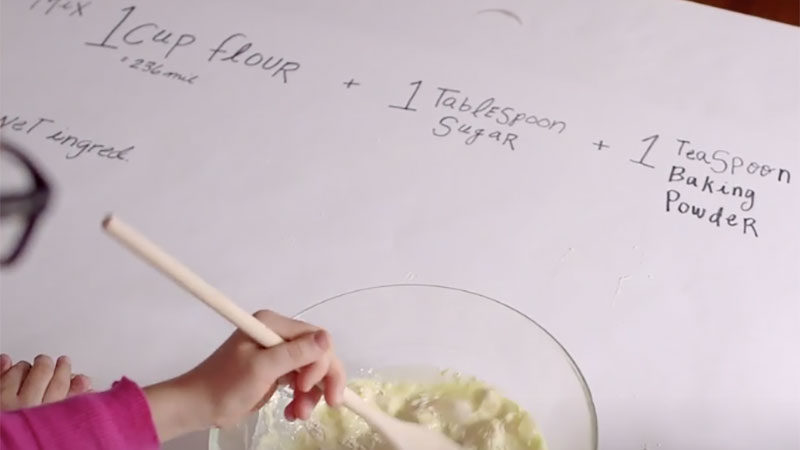 A young girl mixes up pancake ingredients on top of a large sheet of paper with the recipe and measurements.